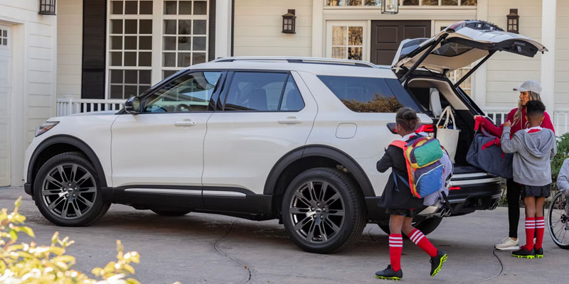 A white Explorer parked in a driveway with two children loading backpacks into the trunk.