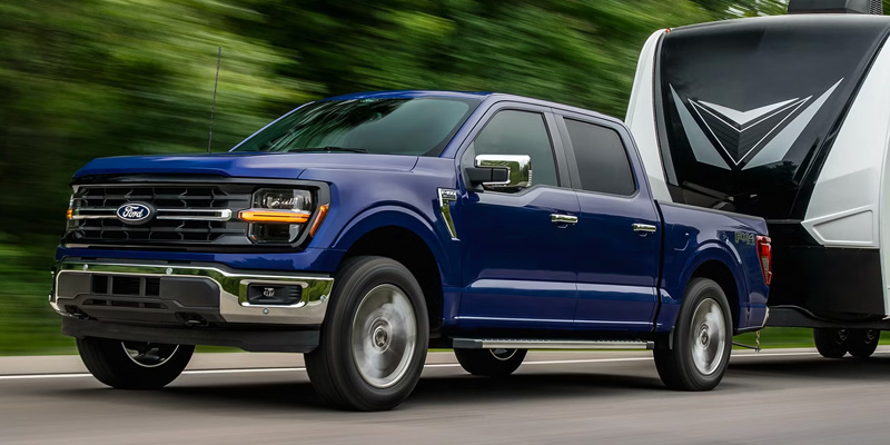 Blue Ford F-150 truck towing a camper on a road surrounded by trees.